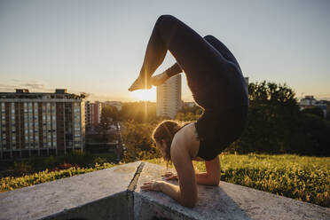 Flexible young woman performing yoga on retaining wall in city during sunset - MEUF01330
