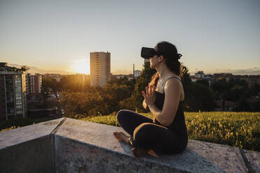Entspannte Frau mit VR-Brille beim Yoga in Gebetshaltung auf einer Stützmauer in der Stadt - MEUF01332