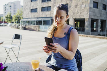 Sportliche junge Frau, die ihr Smartphone benutzt, während sie in einem Straßencafé in der Stadt sitzt - MEUF01336