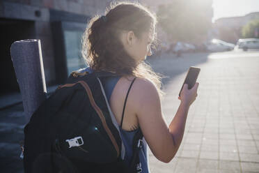 Sportliche junge Frau, die ihr Smartphone benutzt, während sie an einem sonnigen Tag durch die Stadt läuft - MEUF01337