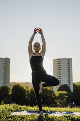 Young woman practicing yoga in tree pose at city park against clear sky - MEUF01342