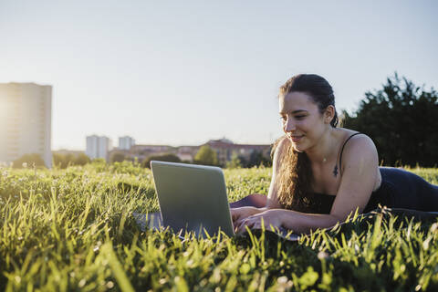 Sporty woman using laptop while lying on grass at city park during sunny day stock photo
