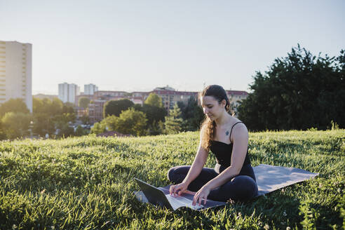 Sportliche junge Frau benutzt einen Laptop, während sie auf einer Übungsmatte im Stadtpark sitzt - MEUF01345