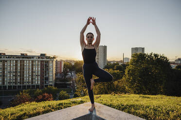 Junge Frau übt Yoga in Baumhaltung auf einer Stützmauer in der Stadt bei Sonnenuntergang - MEUF01347