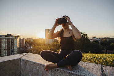 Sportliche Frau, die eine VR-Brille benutzt, während sie bei Sonnenuntergang auf einer Stützmauer in der Stadt sitzt - MEUF01348
