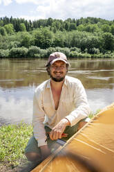 Lächelnder Mann mit Mütze beim Aufbau des Zeltes am See auf einem Campingplatz im Wald - VPIF02612