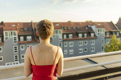 Frau mit kurzen Haaren, die während des Sonnenuntergangs auf einem Balkon steht und das Gebäude betrachtet - TAMF02523