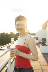 Lächelnde Frau mit Bierflasche auf einem Balkon gegen den klaren Himmel bei Sonnenuntergang - TAMF02525