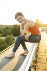 Schöne Frau, die eine Bierflasche hält, während sie auf dem Geländer eines Balkons gegen den klaren Himmel bei Sonnenuntergang sitzt - TAMF02528