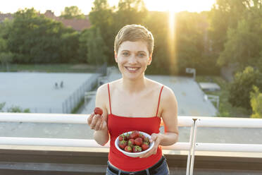 Smiling beautiful woman eating strawberries while standing in balcony at sunset - TAMF02530