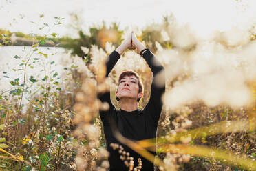 Woman with eyes closed and arms raised exercising while standing amidst plants - MRRF00122