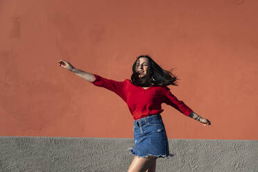 Carefree young woman dancing against orange wall in city on sunny day - MCVF00521