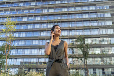 Lächelnde Geschäftsfrau, die über einen drahtlosen Kopfhörer spricht, während sie an einem Gebäude in der Stadt steht - AFVF06723