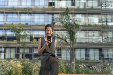 Smiling female entrepreneur using mobile phone while standing against building in city - AFVF06725