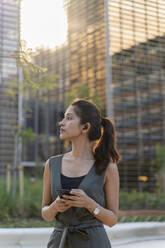 Nachdenkliche Geschäftsfrau hält ihr Smartphone in der Hand, während sie vor modernen Gebäuden in der Stadt steht - AFVF06727