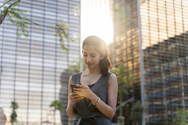 Geschäftsfrau, die ein Smartphone benutzt, während sie vor modernen Gebäuden in der Stadt steht - AFVF06728