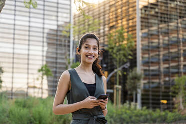 Lächelnde Geschäftsfrau, die ein Mobiltelefon vor modernen Gebäuden in der Stadt benutzt - AFVF06732