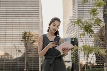 Businesswoman holding note pad using smart phone while standing against modern buildings in city - AFVF06742