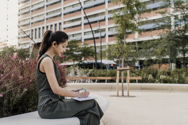 Eine Unternehmerin schreibt in einen Notizblock, während sie auf einem Stuhl vor einem Gebäude sitzt - AFVF06751
