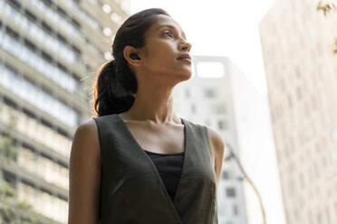 Thoughtful young woman wearing sports bra while listening music against  buildings in city stock photo