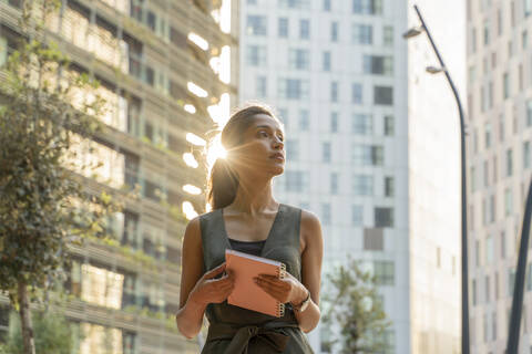 Thoughtful female entrepreneur holding note pad while standing against buildings in modern city stock photo