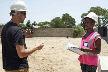 Coworkers wearing helmets discussing while standing at construction site during sunny day - VEGF02496