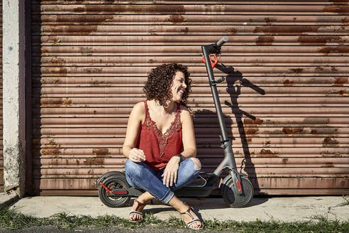 Smiling woman with curly hair sitting on electric push scooter against closed shutter - VEGF02508