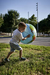Carefree boy holding globe while running on grassy land during sunny day - VEGF02514