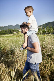 Vater trägt Sohn auf den Schultern beim Spaziergang inmitten von Pflanzen gegen den klaren Himmel an einem sonnigen Tag - VEGF02518