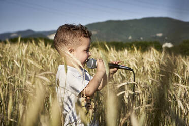 Süßer Junge singt über Mikrofon, während er inmitten von Feldfrüchten an einem sonnigen Tag steht - VEGF02522