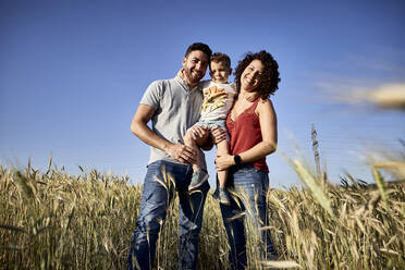 Smiling parents carrying son while standing amidst crops against clear blue sky - VEGF02527