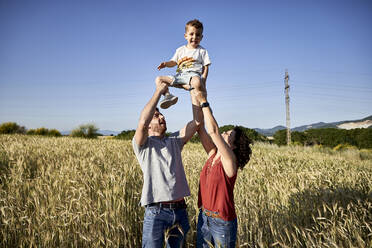 Verspielte Eltern, die ihren Sohn abholen, während sie inmitten von Feldfrüchten vor einem klaren blauen Himmel stehen - VEGF02528