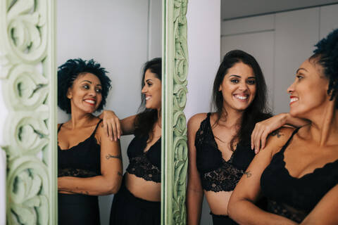 Happy female friends wearing black clothing standing by mirror at home stock photo