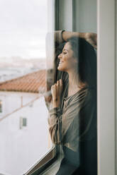 Lächelnde junge Frau, die durch das Fenster ihres Hauses schaut, gesehen durch eine Glastür - DCRF00391