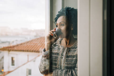 Woman drinking coffee while looking through window at home seen through glass door stock photo