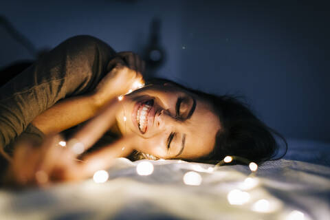 Cheerful young woman with illuminated string lights lying on bed at home stock photo