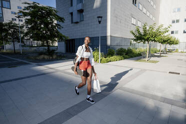 Junge Frau mit Einkaufstasche und Skateboard auf dem Fußweg in der Stadt - MEUF01381