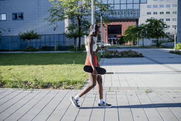 Junge Frau, die ein Skateboard hält und ein Smartphone benutzt, während sie auf einem Fußweg in der Stadt spazieren geht - MEUF01382