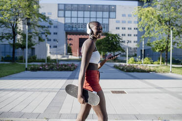 Glückliche Frau, die ein Skateboard hält und ein Mobiltelefon benutzt, während sie auf einem Fußweg in der Stadt spazieren geht - MEUF01384