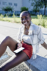 Lächelnde junge Frau mit rasiertem Kopf, die ein Mobiltelefon benutzt, während sie auf einem Fußweg in der Stadt sitzt - MEUF01385