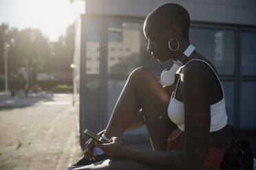 Junge Frau mit rasiertem Kopf, die ein Smartphone benutzt, während sie an einem sonnigen Tag in der Stadt sitzt - MEUF01389