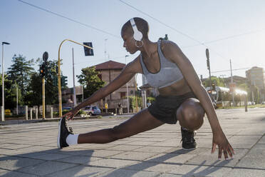 Young woman listening music while exercising on street against clear sky in city - MEUF01400