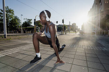 Woman with shaved head listening music through headphones while stretching legs on street in city - MEUF01403
