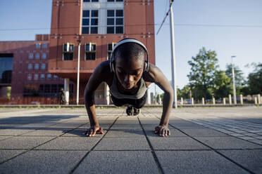 Female athlete with shaved head listening music through headphones while doing push-ups on street in city - MEUF01407