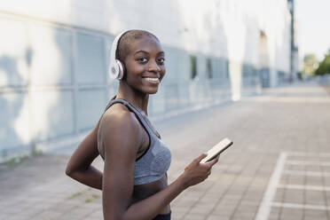 Smiling young woman wearing headphones using smart phone while standing in city - MEUF01412