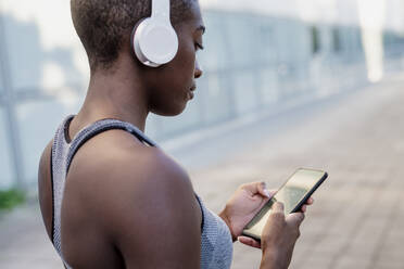 Nahaufnahme einer jungen Frau mit Kopfhörern, die ein Smartphone benutzt, während sie in der Stadt steht - MEUF01413