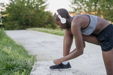 Female athlete wearing headphones tying shoelace on retaining wall in park - MEUF01422