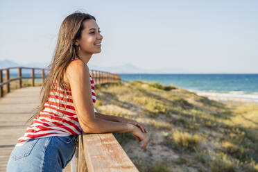 Thoughtful teenage girl standing on boardwalk at beach against clear blue sky - DLTSF00856