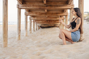 Glückliches Teenager-Mädchen nimmt Selfie durch Smartphone unter Pier am Strand - DLTSF00857