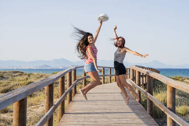 Aufgeregte Teenager-Mädchen springen auf der Promenade am Strand gegen den klaren Himmel - DLTSF00863